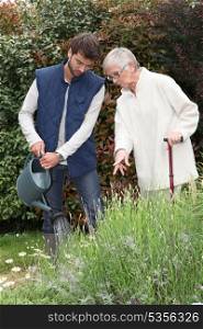 A gardener watering flowers in a garden and an elderly lady making comments as she watches him.