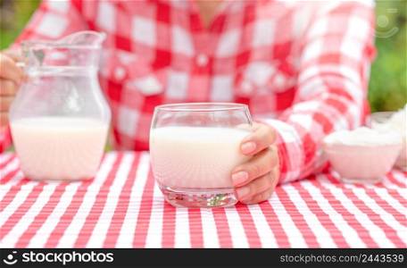 A girl in a red plaid shirt holds out a glass of milk. Summer sunny day. Next to the table are dairy products. Country landscape. Natural green background. The concept of natural rustic milk.. A girl in a red plaid shirt holds out a glass of milk