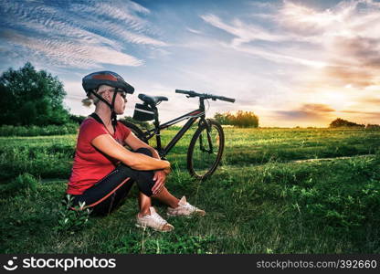 A girl in bicycle helmet and glasses sits near bicycle and looks at the sunset. Summer landscape. The concept of freedom and travel.. A girl in bicycle helmet sits near bicycle