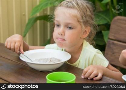 A girl of four years at a table outdoors thoughtfully eating porridge