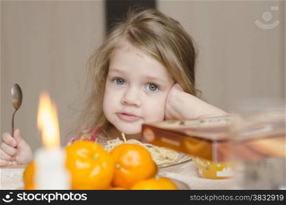 A girl of two years sits with a spoon at the festive table and eats macaroni