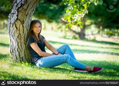A girl sits under a tree in a sunny park and looks into the frame a. A girl sits under a tree in a sunny park and looks into the frame