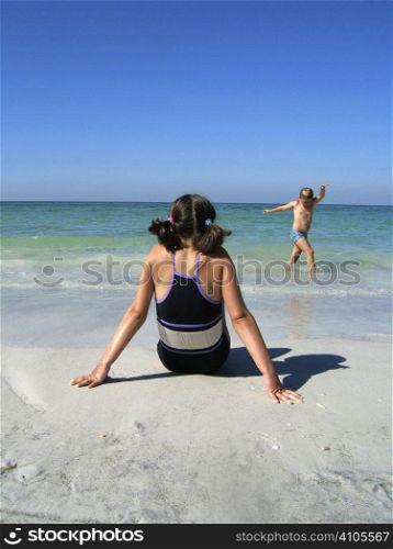 a girl sitting on the beach while her brother plays in the sea