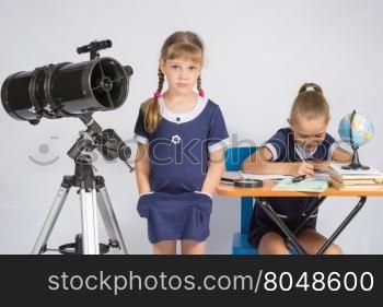 A girl stands at a telescope with his hands in his pockets, another girl having fun sitting at the table and writes