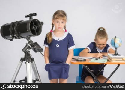 A girl stands at a telescope with his hands in his pockets, another girl having fun sitting at the table and writes