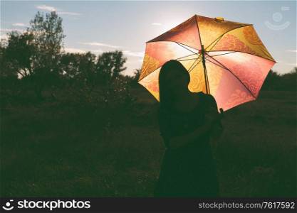 A girl with a colored umbrella poses in a field. The umbrella is illuminated from behind by the sunset light. There&rsquo;s room on the left for a copyspace.. A girl with a colored umbrella poses in a field. The umbrella is illuminated from behind by the sunset light