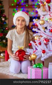 A girl with a red ball sits by the Christmas tree, in the background are blurry flashlights