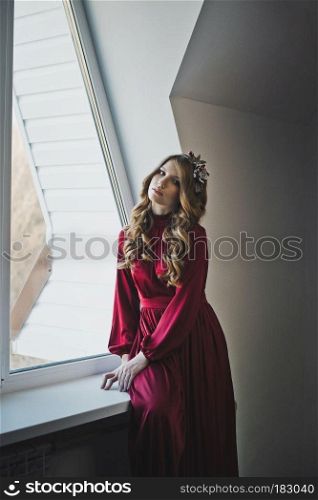 A girl with long brown hair by the window.. Portrait of a girl in a red dress at the window 4424.