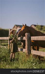 A girl with red hair strokes and feeds grass to horses.. A girl in a green dress is stroking and feeding horses 3243.