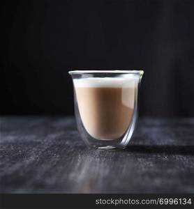 A glass cup of hot flavored cappuccino presented on a black wooden table with copy space.. Freshly brewed cappuccino with foam in a glass cup on a black wooden table around a dark background with copy space.