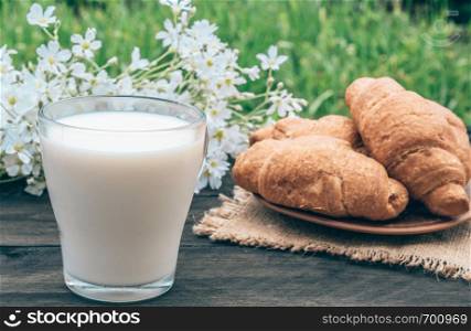A glass cup of milk stands beside croissants and white small flowers.. Next to the croissants and white flowers is a glass cup of milk.