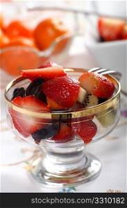 A glass dessert bowl with fresh fruit salad in it, on a tablecloth. Other fruits are visible in the background