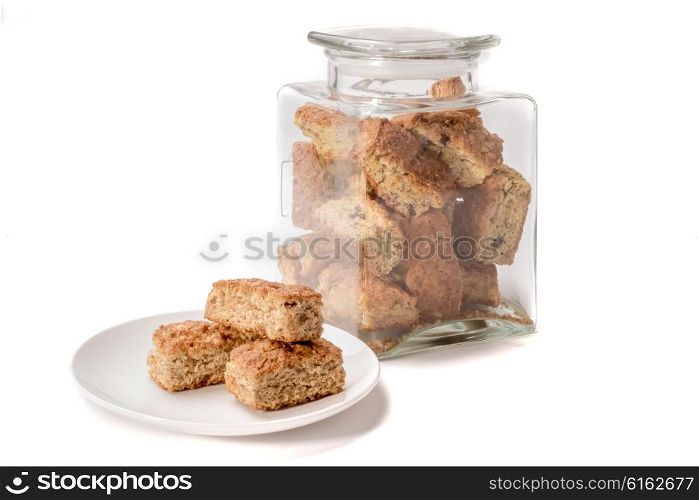 A glass jar filled with golden brown rusks stand behind a white plate with three pieces of rusks stacked, all on a white background.