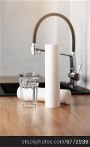A glass of clean water and foamed polypropylene filter cartridges on wooden table in a kitchen interior. Installation of reverse osmosis water purification system. Concept Household filtration system. A glass of clean water and foamed polypropylene filter cartridges on wooden table in a kitchen interior. Installation of reverse osmosis water purification system. Concept Household filtration system.