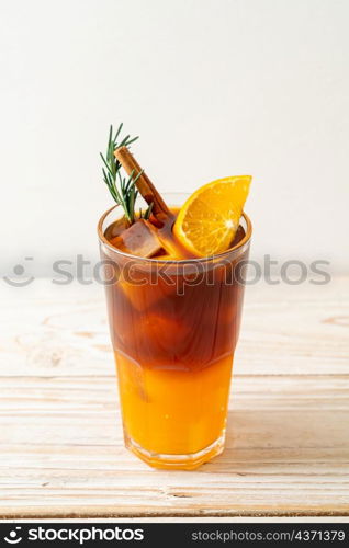 A glass of iced americano black coffee and layer of orange and lemon juice decorated with rosemary and cinnamon on wood background