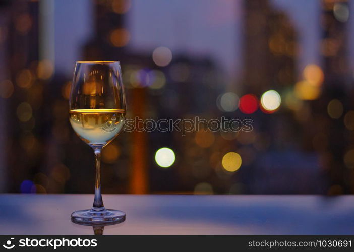 A glass of white wine on table of rooftop bar with colorful bokeh of city light and space for text.