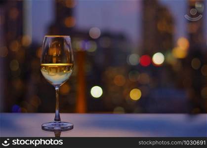 A glass of white wine on table of rooftop bar with colorful bokeh of city light and space for text.