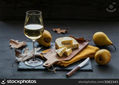 A glass of white wine was served with cheese in a cutting board on dark background. Autumn picnic with cheese, wine and dry leaves in rustic style.