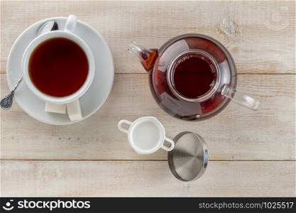 a glass teapot, mug of hot black tea and a milk jug on a wooden table. a glass kettle, tea cup of hot black tea and a milk jug on a wooden table.Top view