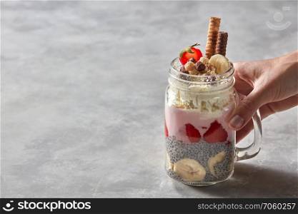 A glass with a handle in a woman’s hand with a healthy breakfast of yogurt, strawberry, chia pudding with a banana, decorated with biscuits and whipped cream on a gray concrete background. Copy space. Glass with a layered breakfast from yogurt, strawberry, chia pudding with a banana in the female hand on stone background