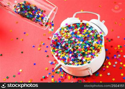 A glass with multicolored sequins, alarm clock and star shaped confettie on the red background. Party, new year, celebration concept. A glass with multicolored sequins, alarm clock and star shaped confettie on the red background. Party, new year, concept