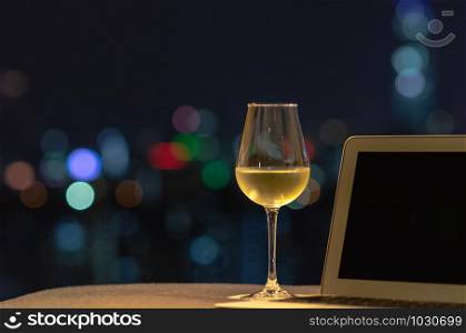 A glass with white wine puts on sofa with laptop computer with colorful bokeh light from city.