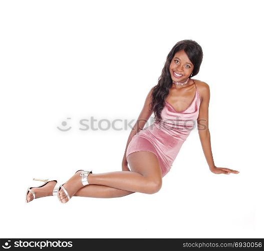 A gorgeous young African woman sitting in a short pink dress on thefloor, smiling, isolated for white background