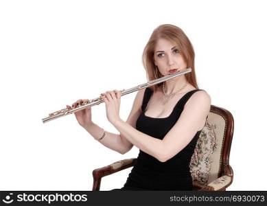 A gorgeous young woman sitting in a black dress and playing theflute, looking into the camera, isolated for white background