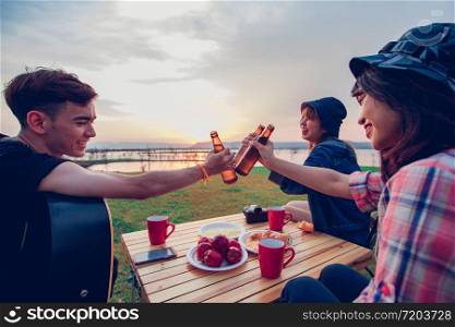 A group of Asian friends tourist drinking alcoholic beer and playing guitar together with happiness in Summer while having camping near lake at sunset