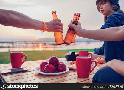 A group of Asian friends tourist drinking alcoholic beer and playing guitar together with happiness in Summer while having camping near lake at sunset