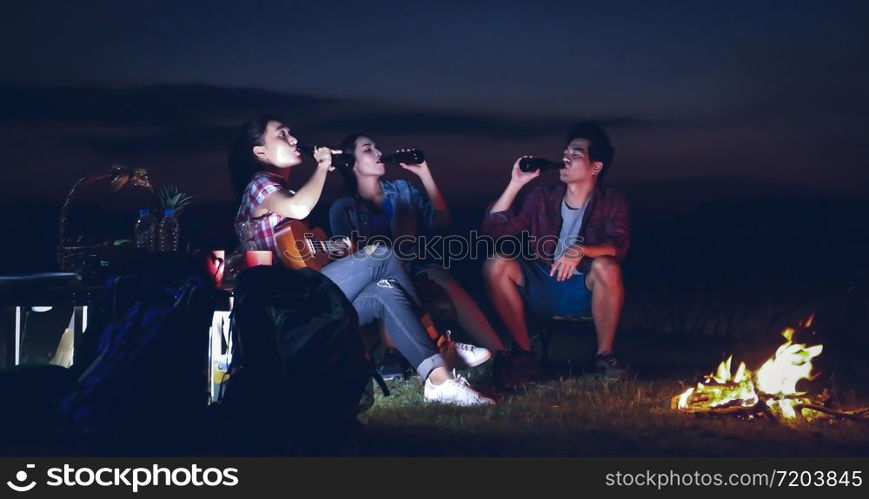 A group of Asian friends tourist drinking and playing guitar together with happiness in Summer while having camping near lake at sunset