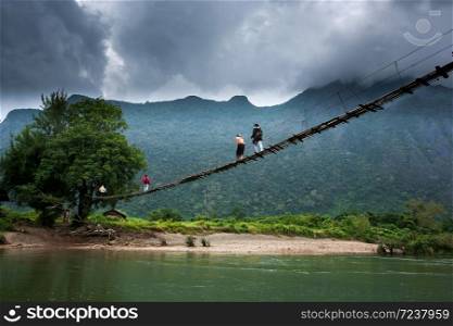 A group of Laotian high school girls across the simple suspension footbridge over the Nam Song River, dramatic dark cloudy over mountain in the backgrounds, rural scene in North Laos.