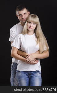 A half-length portrait of a young couple. Guy hugging a girl back. Studio, black background.