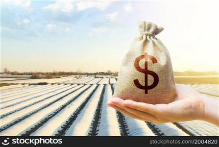 A hand holds out a dollar money bag on a background of white spunbond agrofibre rows on a farm field. Lending farmers and agricultural enterprises. Support and subsidies. Protective coating for corps.