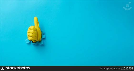 a hand in a yellow household rubber glove sticks out of a torn hole and shows a gesture okay, thumb up. Blue background
