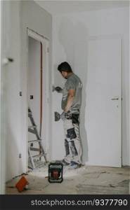 A handsome young Caucasian man in a gray uniform and gloves standing puttying a corner in a doorway with spatulas in the evening with a building lantern on, close-up side view. Construction concept.. A young builder is plastering a doorway.