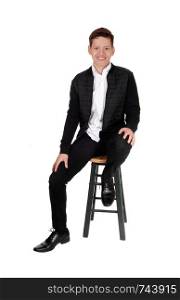A handsome youth teen boy sitting on a chair in the studio is a black jacket and pants, smiling, isolated for white background