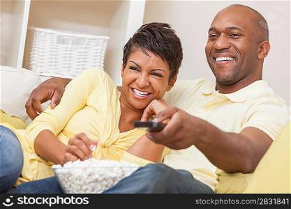A happy African American man and woman couple in their thirties sitting at home, eating popcorn and using remote control watching a movie or television