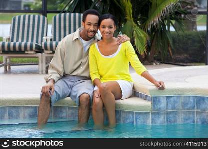A happy African American man and woman couple in their thirties sitting with their feet in a swimming pool.