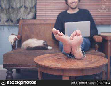A happy barefoot man is resting his feet on a coffee table at home while working on his laptop, there is a cat sleeping next to him