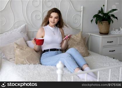 A happy beautiful business consultant woman in formal attire, using a mobile phone, reading the news or sending text messages, sitting in a room with a bed, enjoying a busy working day. Cute girl is sitting on a white bed with a phone and a red mug