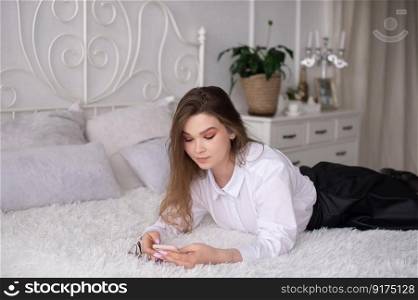 A happy beautiful business consultant woman in formal attire, using a mobile phone, reading the news or sending text messages, sitting in a room with a bed, enjoying a busy working day. A young business woman is lying on her bed and reading the news or sending a text message.