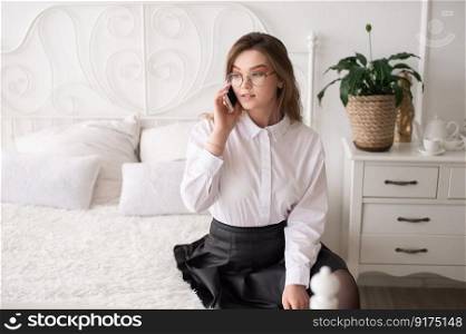 A happy beautiful business consultant woman in formal attire, using a mobile phone, reading the news or sending text messages, sitting in a room with a bed, enjoying a busy working day. A happy emotional European girl in a white shirt and glasses is talking on the phone. Sitting on a white bed