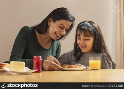 A HAPPY DAUGHTER SITTING WITH MOTHER AND SPREADING JAM ON BREAD
