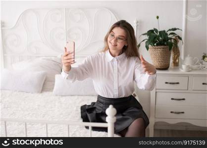 A happy European woman has a video call on her smartphone . The girl looks into the phone camera and smiles. White Scandinavian interior. Daylight through the window.. Weekend morning at home. Emotional Caucasian woman talking on the phone