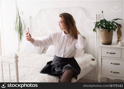 A happy European woman has a video call on her smartphone . The girl looks into the phone camera and smiles. White Scandinavian interior. Daylight through the window.. An emotional successful European woman with a phone straightens her hair for a selfie.