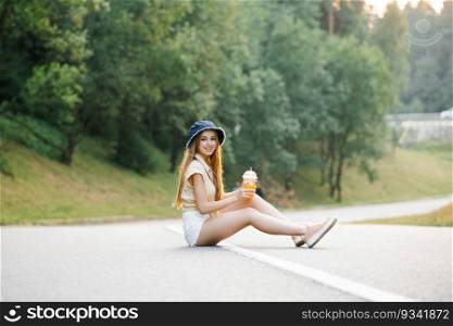 A happy girl in white shorts and a yellow blouse holds a glass of juice with a straw in her hands and sits on the road near the forest