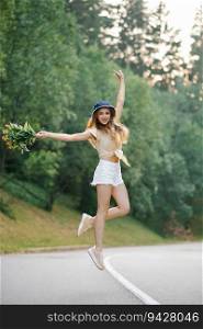 A happy girl in white shorts and a yellow blouse jumps up with a bouquet of flowers, standing on the road near the forest