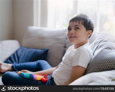 A happy kid sitting on a sofa playing a multi-colored plastic toy popit. Indoor portrait with a cute child relaxing and enjoy playing with toys at home, New Fidget toy is popular for Kids