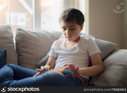 A happy kid sitting on a sofa playing a multi-colored plastic toy popit. Indoor portrait with a cute child relaxing and enjoy playing with toys at home, New Fidget toy is popular for Kids